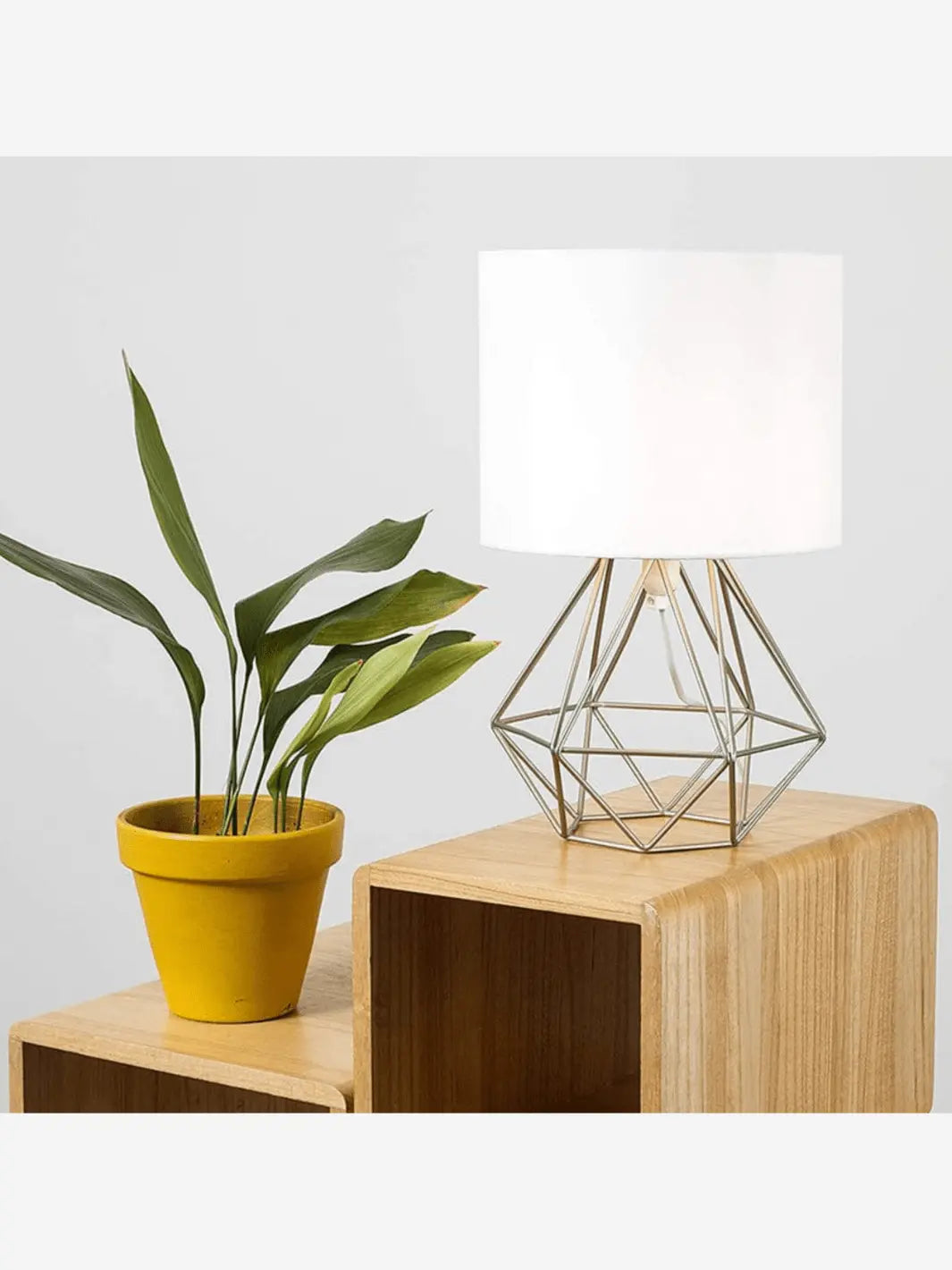 lampe de chevet metal moderne au design avec un pied geometrique couleur argent et abat jour blanc posee sur un meuble design en bois a cote d une plante